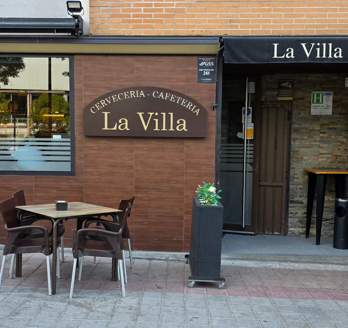 Entrada de La Villa: rótulo de madera con letras doradas «Cervecería - Cafetería La Villa», toldo negro con el nombre y una mesa en la acera