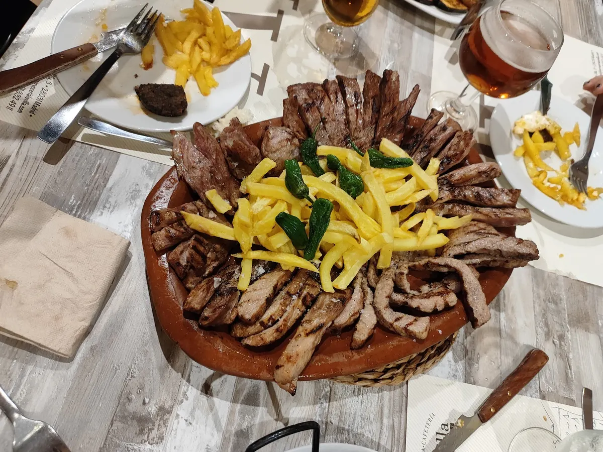 Fuente de barro con carnes a la parrilla, patatas fritas y pimientos del padrón, con dos cañas de cerveza en la mesa