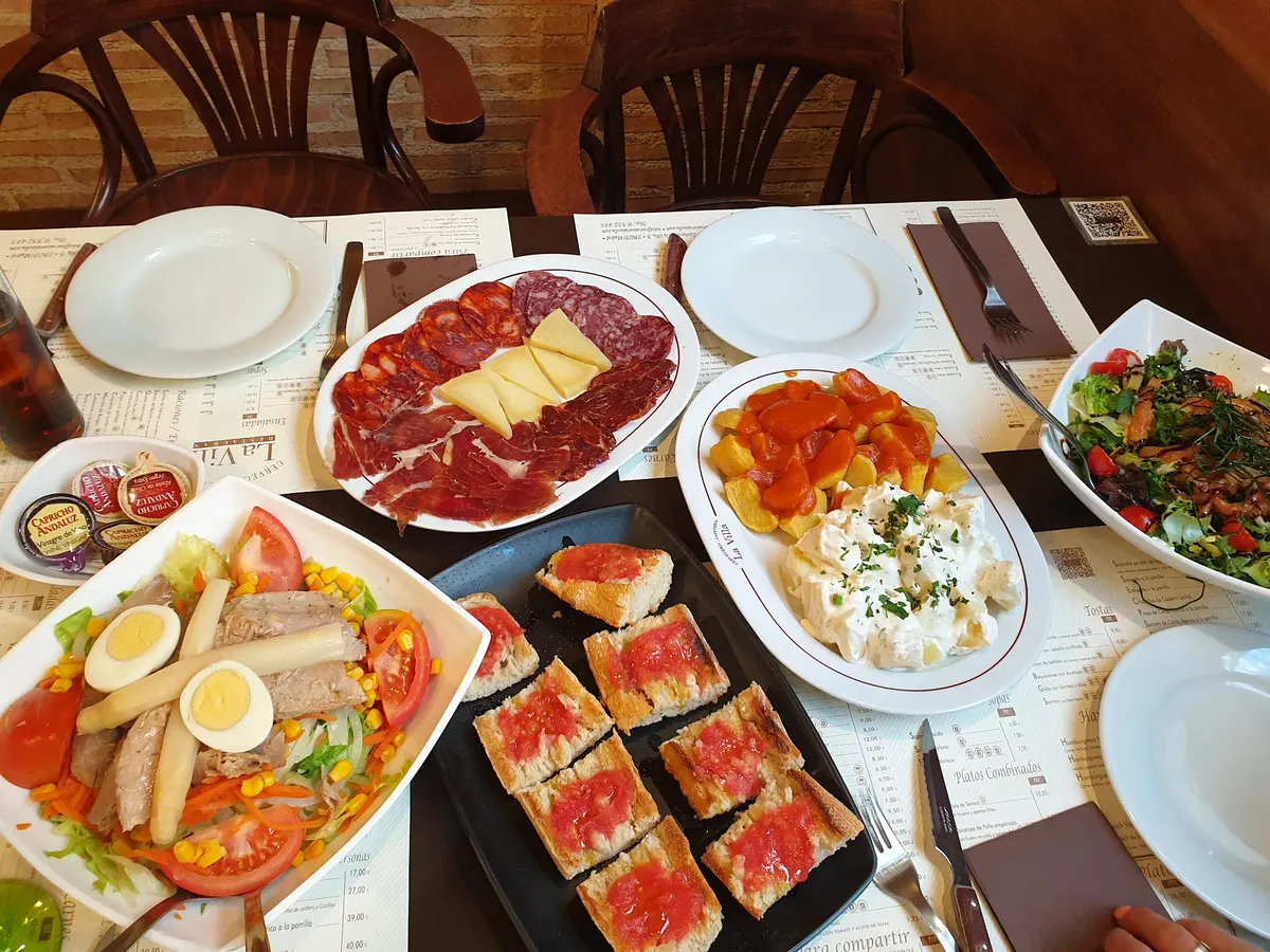 Mesa con raciones para compartir: surtido de ibéricos con queso, patatas bravas, ensaladilla, pan tostado con tomate y dos ensaladas