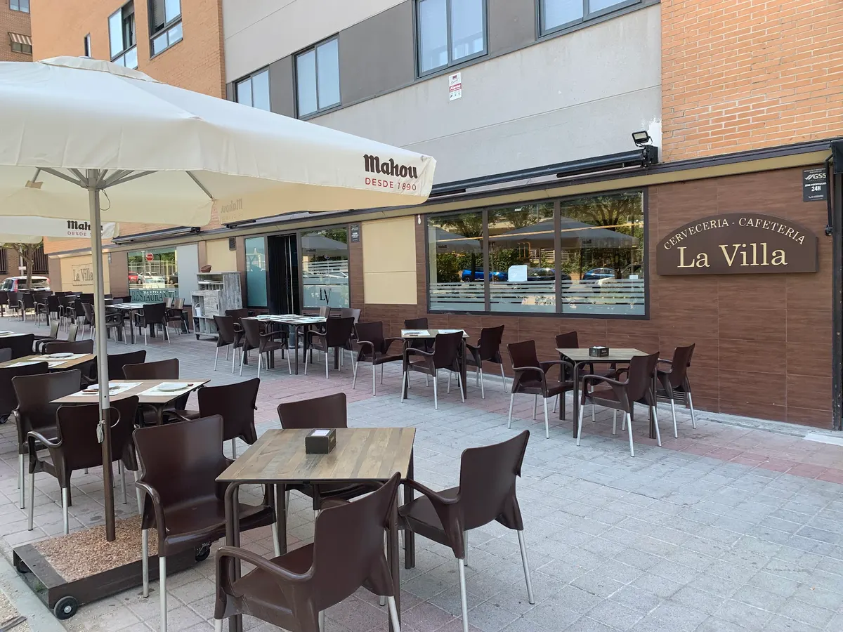 Terraza de La Villa con mesas de madera, sillas marrones y sombrillas, junto a la fachada de ladrillo con el rótulo «Cervecería - Cafetería La Villa»